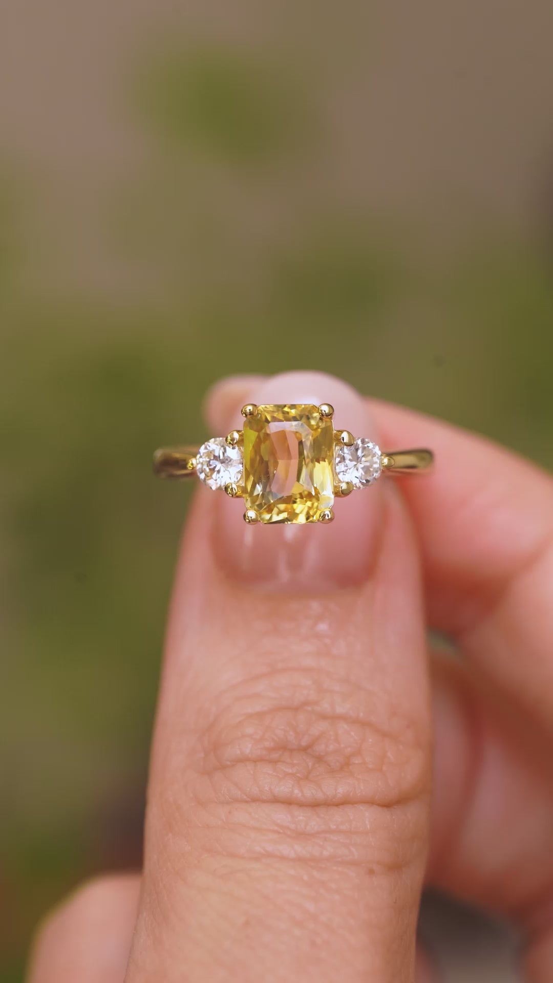 A video of a vintage style, Yellow sapphire and diamond three-stone engagement ring in yellow gold held between the fingers against a blurry background.