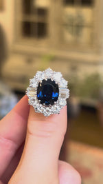 Large oval 5ct sapphire and diamond cluster ring held between fingers.