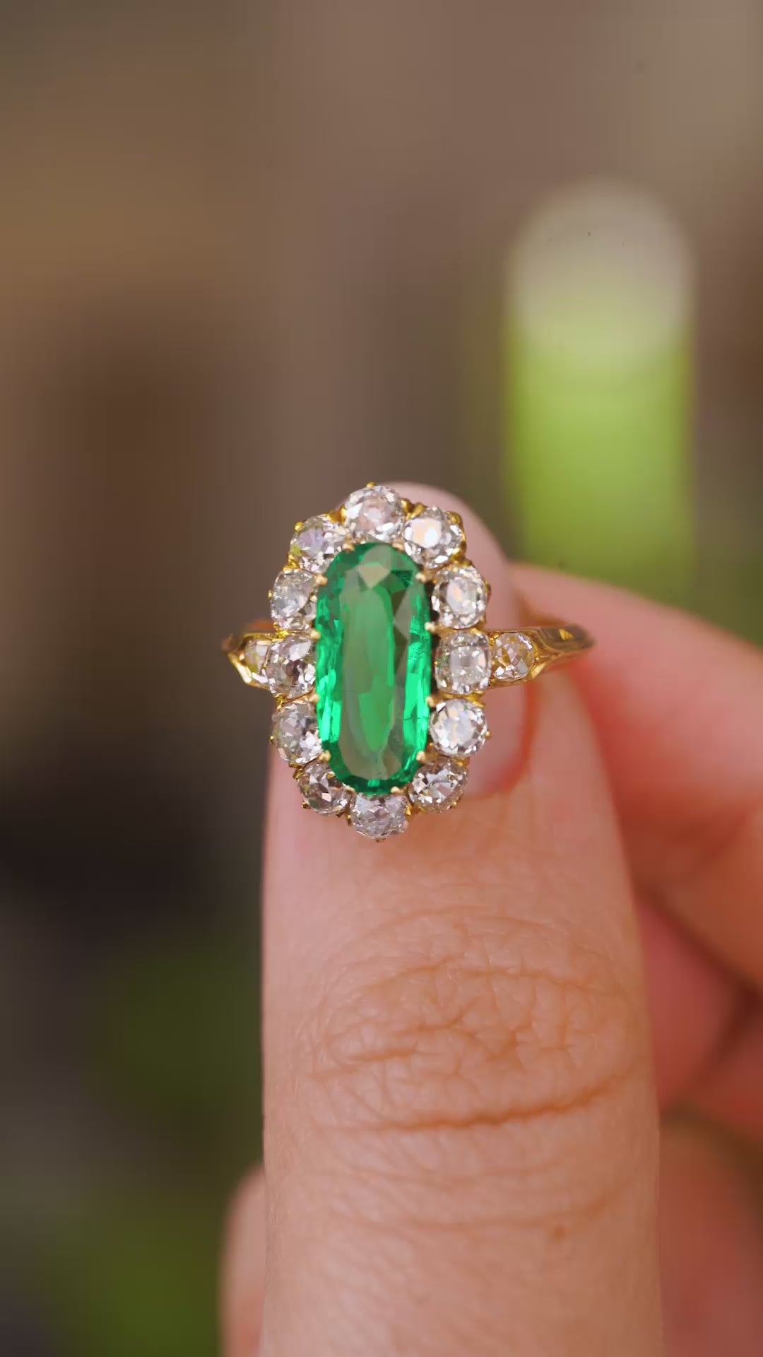 A close up video of an Antique, Belle Époque emerald and diamond cluster engagement ring.  Held between two fingers and rotated slowly to showcase the elongated oval of the central Colombian emerald and the yellow gold band.  