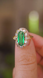 A close up video of an Antique, Belle Époque emerald and diamond cluster engagement ring.  Held between two fingers and rotated slowly to showcase the elongated oval of the central Colombian emerald and the yellow gold band.  