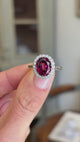 Vintage 1960s garnet and diamond cluster engagement ring showcasing an oval garnet centre stone and diamond halo, held by hand to highlight the setting and craftsmanship.