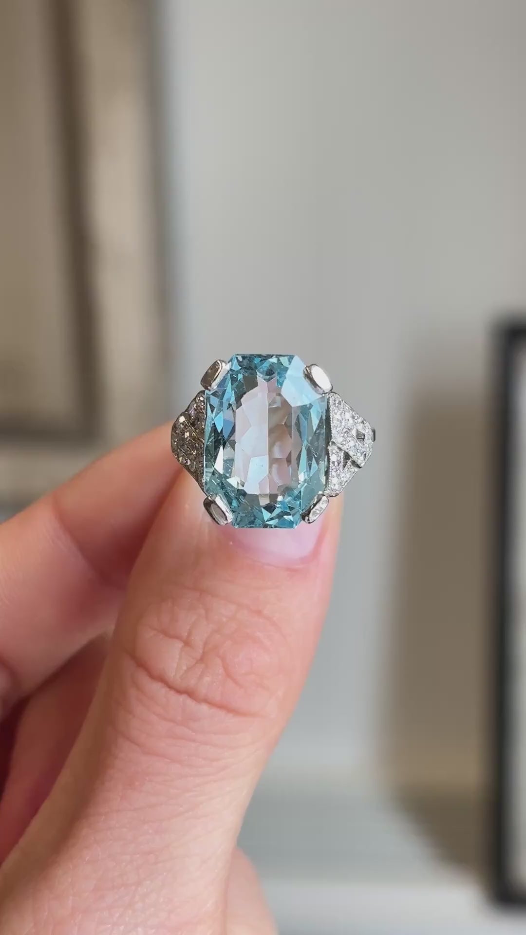 Close-up video of an antique Art Deco aquamarine and diamond cocktail ring in platinum, highlighting the large faceted aquamarine, diamond surround and elegant craftsmanship against a softly blurred antique-inspired background.