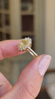 An Antique-inspired, vibrant yellow sapphire and diamond cluster engagement ring in yellow gold, held between 2 fingers with a wedding band to show the profile and wedding fit of the ring.