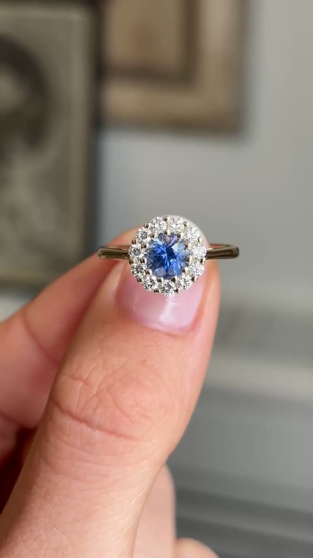 Close up video of an Antique-inspired, electric blue sapphire and diamond cluster engagement ring in yellow gold, held between 2 fingers and rotated to show the side profiles of the ring. 