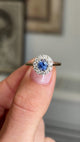 Close up video of an Antique-inspired, electric blue sapphire and diamond cluster engagement ring in yellow gold, held between 2 fingers and rotated to show the side profiles of the ring. 