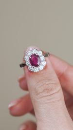A video showing the white gold ruby engagement ring held between fingers, revealing its classic oval cluster design and glittering diamond border.