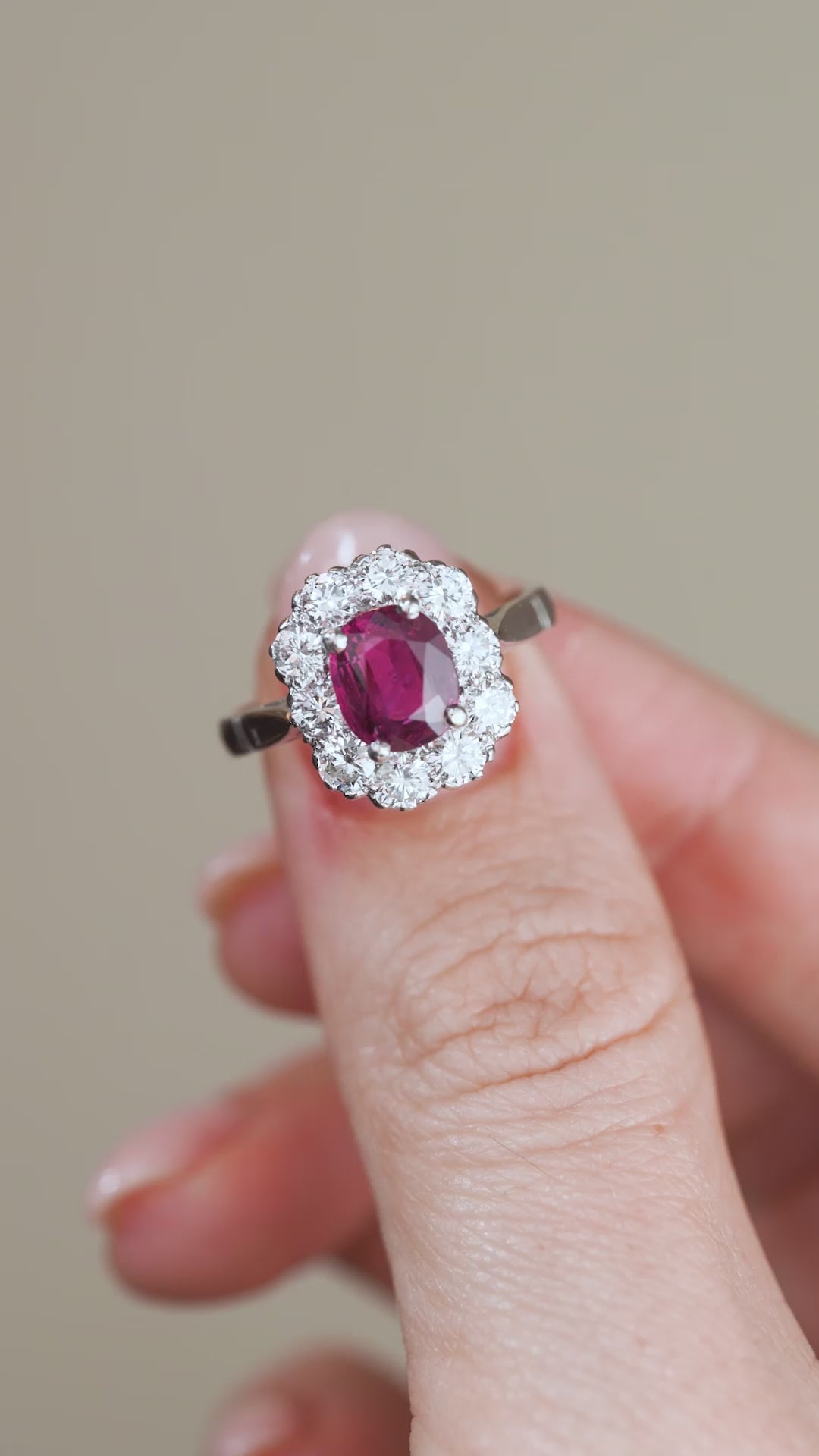A video showing the white gold ruby engagement ring held between fingers, revealing its classic oval cluster design and glittering diamond border.