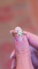 Cushion old-mine cut diamond solitaire engagement ring held between fingers showing ring details in motion.