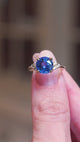 Round blue sapphire and diamond cluster tre-foil engagement ring held between fingers showing ring details in motion.