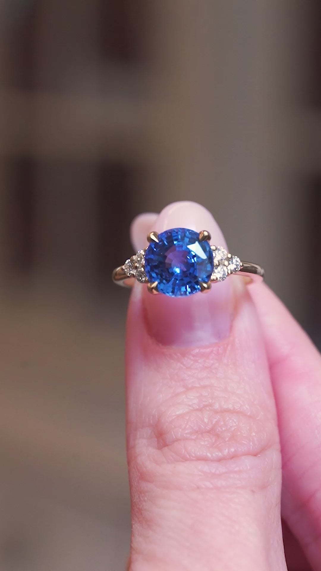 Round blue sapphire and diamond cluster tre-foil engagement ring held between fingers showing ring details in motion.