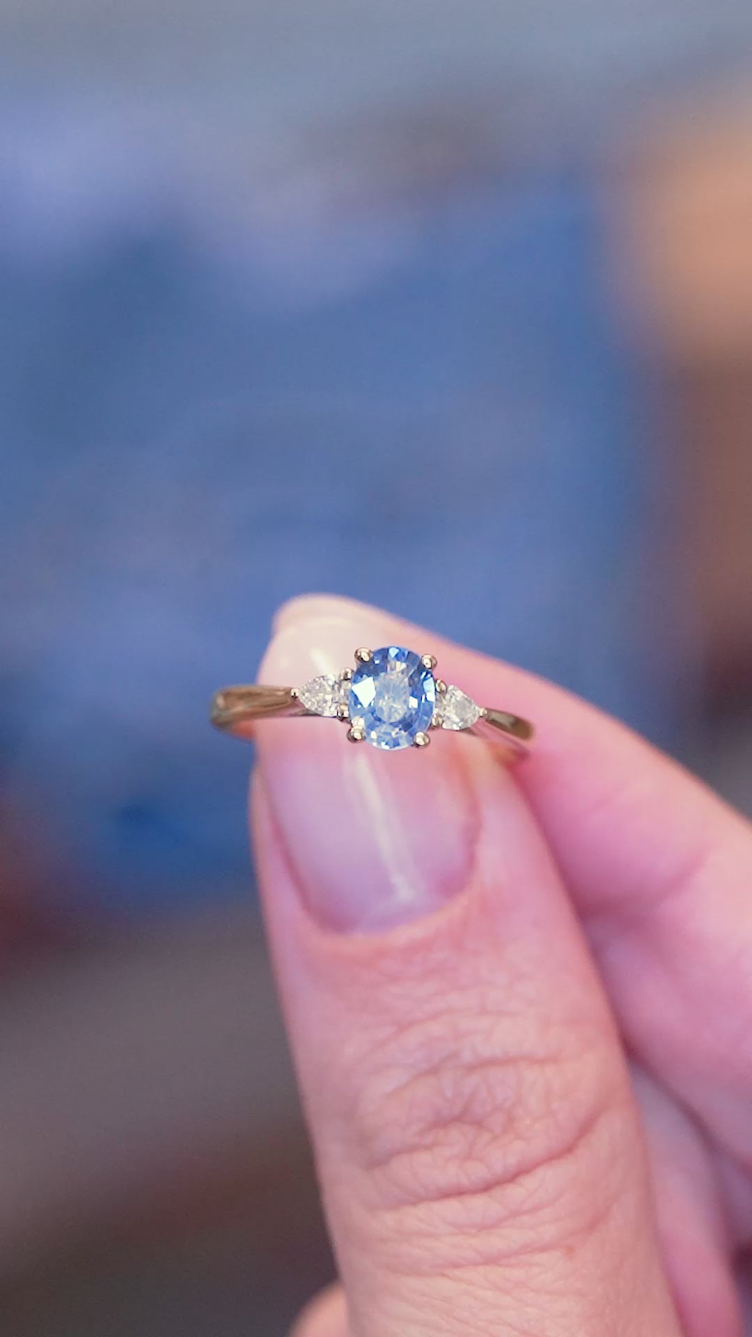 Light blue sapphire and diamond three-stone engagement ring worn held between fingers showing ring details.