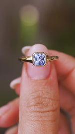 A close up video of an Antique-inspired, blue sapphire solitaire engagement ring held between two fingers and rotated slowly to showcase the yellow gold bezel setting of the Sri Lankan sapphire and the yellow gold band.  