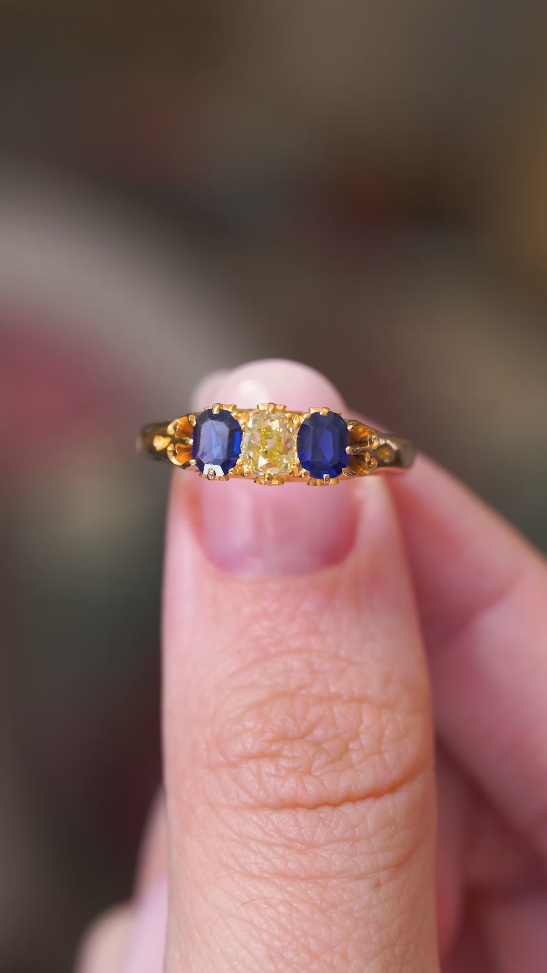 A video of an Antique, Victorian blue sapphire and cushion-cut yellow diamond three-stone engagement ring held between the thumb and finger and rotated to display all of the angles, seen against a blurred background.