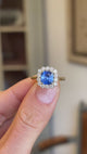 Close-up video of an antique-inspired violet-blue cushion cut Sri Lankan sapphire and diamond cluster engagement ring in yellow gold, highlighting the vibrant cushion cut sapphire, brilliant diamond surround and exceptional sparkle against a softly blurred interior setting.