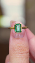 A video of a close-up of an antique-inspired emerald-cut emerald solitaire engagement ring in yellow gold held between fingers.