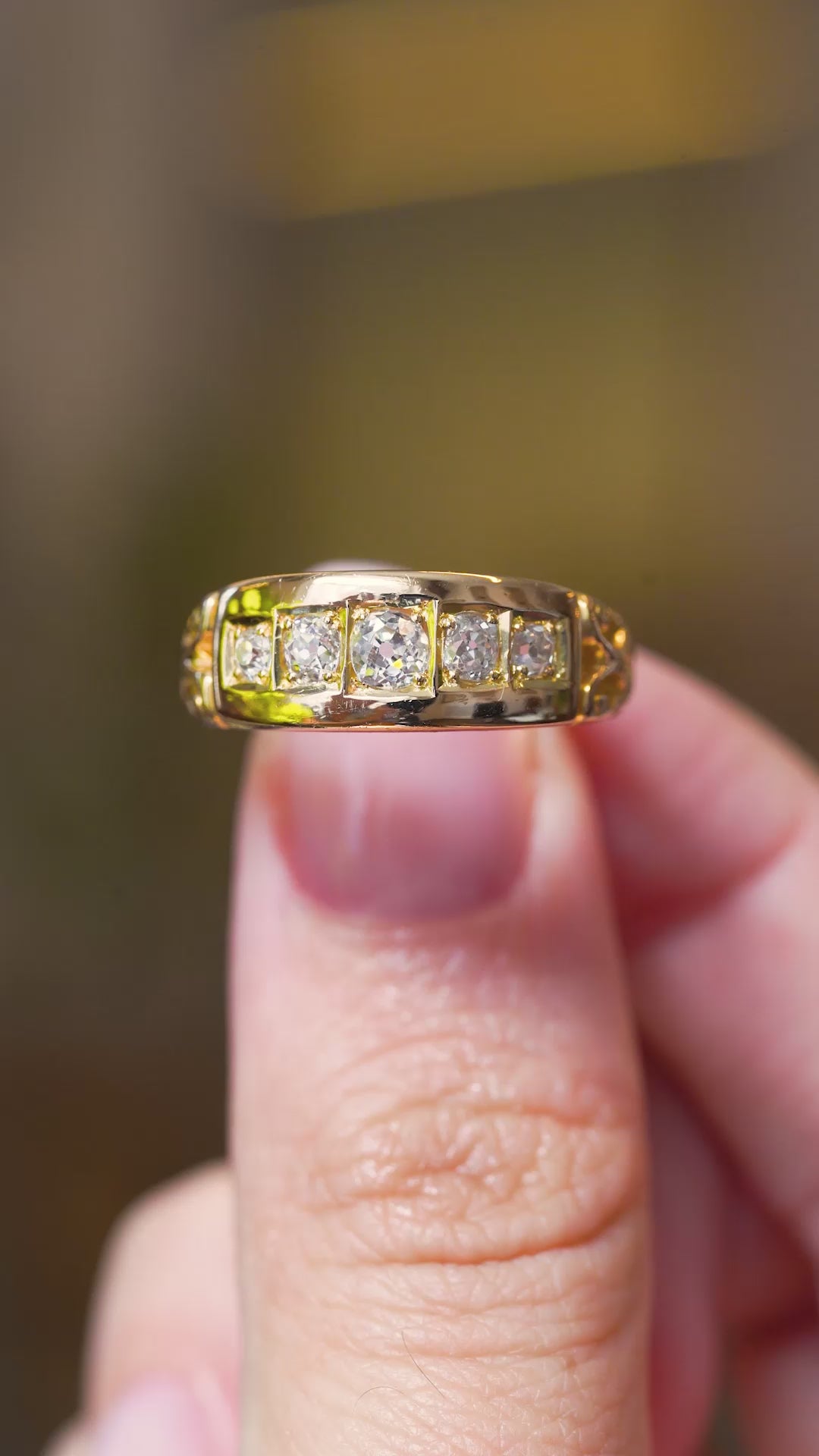 A video of an Antique, Victorian diamond five-stone engagement ring mounted in yellow gold and held between the thumb and the fingers and turned to show off different angles against a blurry background.