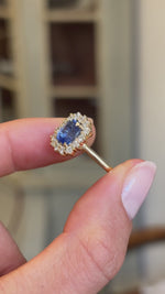 Detailed video of an antique-inspired violet-blue cushion cut Sri Lankan sapphire and diamond cluster engagement ring in yellow gold held between fingers, showcasing the vivid violet-blue sapphire, sparkling diamond halo and fine craftsmanship with a vintage-inspired background.