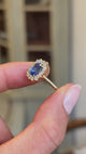 Detailed video of an antique-inspired violet-blue cushion cut Sri Lankan sapphire and diamond cluster engagement ring in yellow gold held between fingers, showcasing the vivid violet-blue sapphire, sparkling diamond halo and fine craftsmanship with a vintage-inspired background.