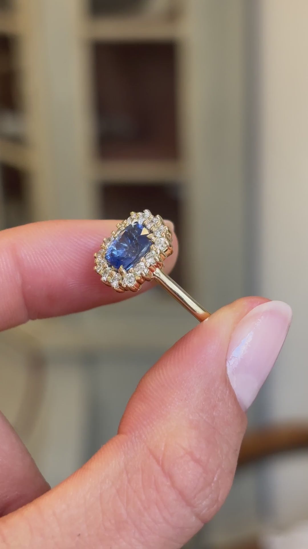 Detailed video of an antique-inspired violet-blue cushion cut Sri Lankan sapphire and diamond cluster engagement ring in yellow gold held between fingers, showcasing the vivid violet-blue sapphire, sparkling diamond halo and fine craftsmanship with a vintage-inspired background.