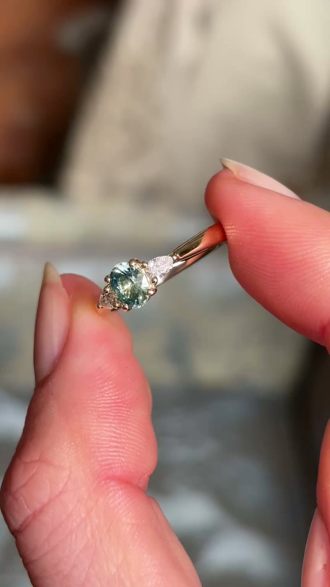 An oval-cut teal sapphire just under 1ct and pear-cut diamond three-stone engagement ring set in yellow gold, between two fingers.