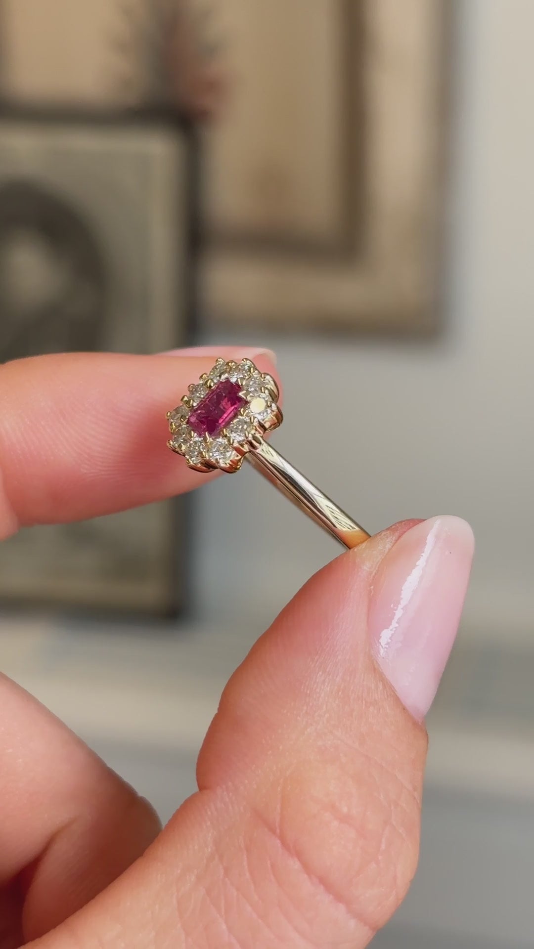 Detailed video of a step-cut pinkish-red ruby and diamond cluster ring in yellow gold, held between fingers to showcase the vivid ruby centre stone, diamond halo and elegant antique-inspired design against a vintage interior setting.