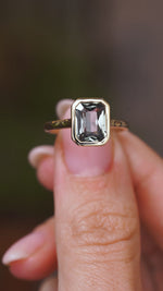 A close up video of an Antique-inspired, grey-blue sapphire solitaire engagement ring.  Held between two fingers and rotated slowly to display the bezel setting of the sapphire and the yellow gold band. 