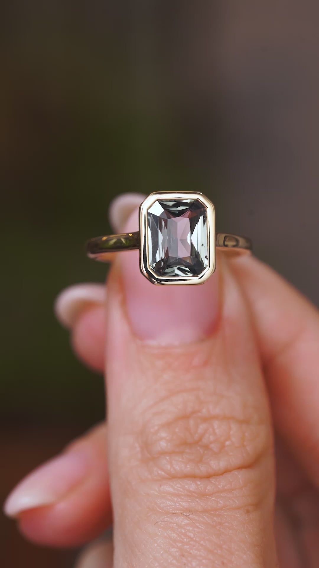 A close up video of an Antique-inspired, grey-blue sapphire solitaire engagement ring.  Held between two fingers and rotated slowly to display the bezel setting of the sapphire and the yellow gold band. 