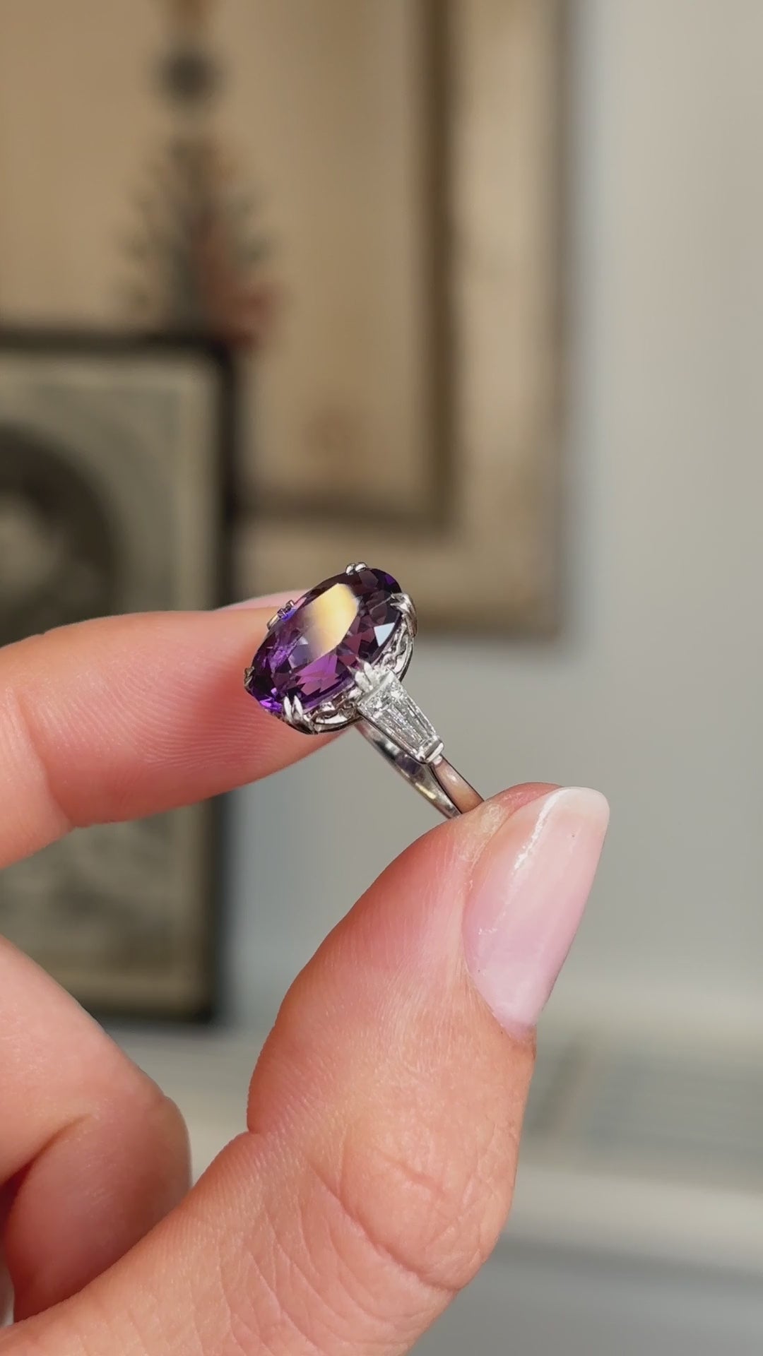 Detailed video of an antique Art Deco amethyst and diamond flanked solitaire three-stone ring in platinum, held between fingers to showcase the purple amethyst centre stone, diamond accents and refined geometric design against a warm interior setting.