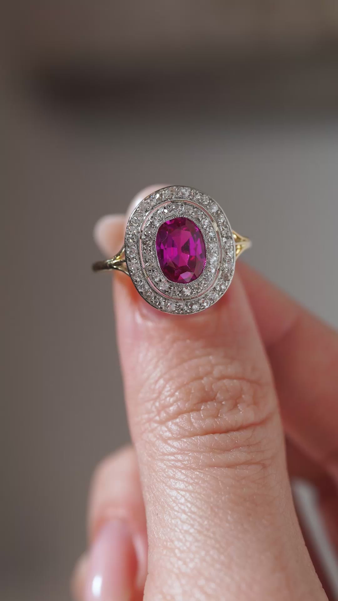 A close-up video of an antique Edwardian Burmese ruby and diamond cluster ring held between fingers, highlighting the ruby's vibrant colour and intricate diamond-set platinum surround.