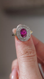 A close-up video of an antique Edwardian Burmese ruby and diamond cluster ring held between fingers, highlighting the ruby's vibrant colour and intricate diamond-set platinum surround.