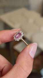 A close-up of a vintage-inspired, pink sapphire and diamond cluster engagement ring held between two fingers shown in video