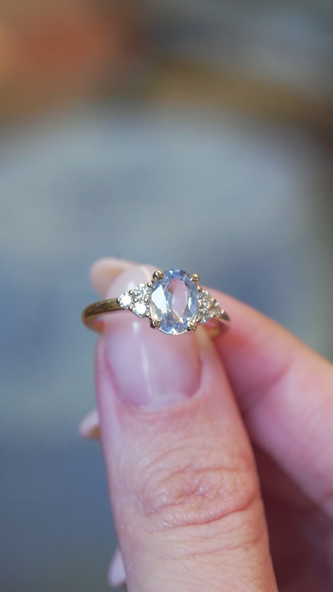 Pale blue sapphire and diamond cluster tre-foil engagement ring held between fingers with motion showcasing the sparkle.