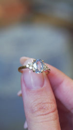 Pale blue sapphire and diamond cluster tre-foil engagement ring held between fingers with motion showcasing the sparkle.