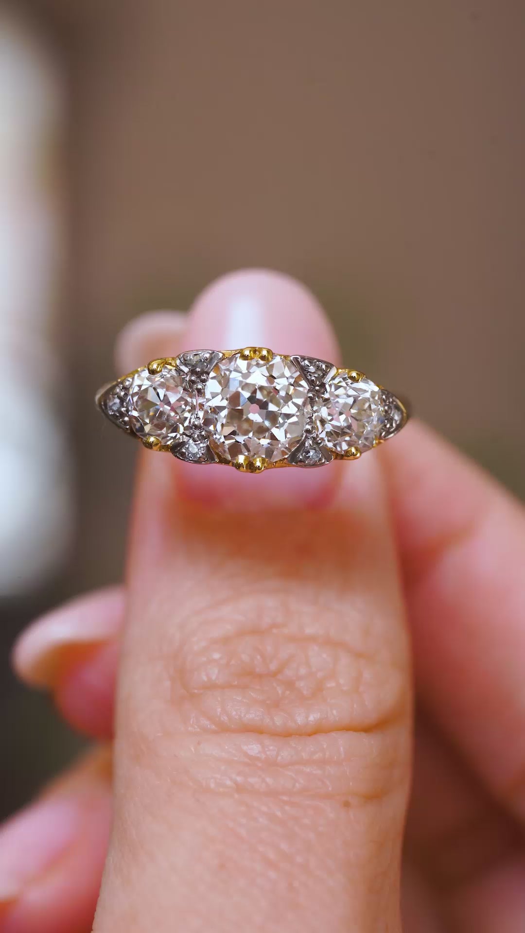 An antique Victorian ring held between two fingers, showing its cushion-shaped old-mine-cut centre diamond flanked by two round old-cut diamonds in a video.