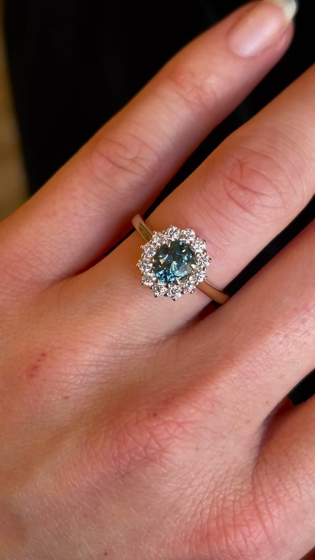 Teal sapphire and diamond cluster yellow gold engagement ring on hand, shown in motion to indicate scale.