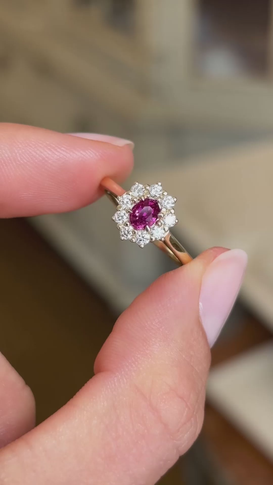 A vintage-inspired engagement ring, with a pink ruby and diamond halo in yellow gold shown between two fingers.