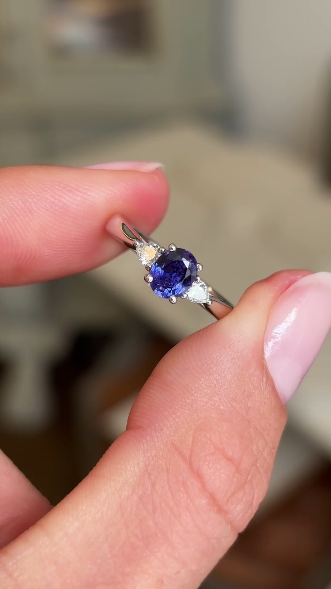 Close up video of a platinum vintage-inspired three stone ring with a central deep indigo blue sapphire flanked by two diamonds.  Held between two fingers against a blurred background