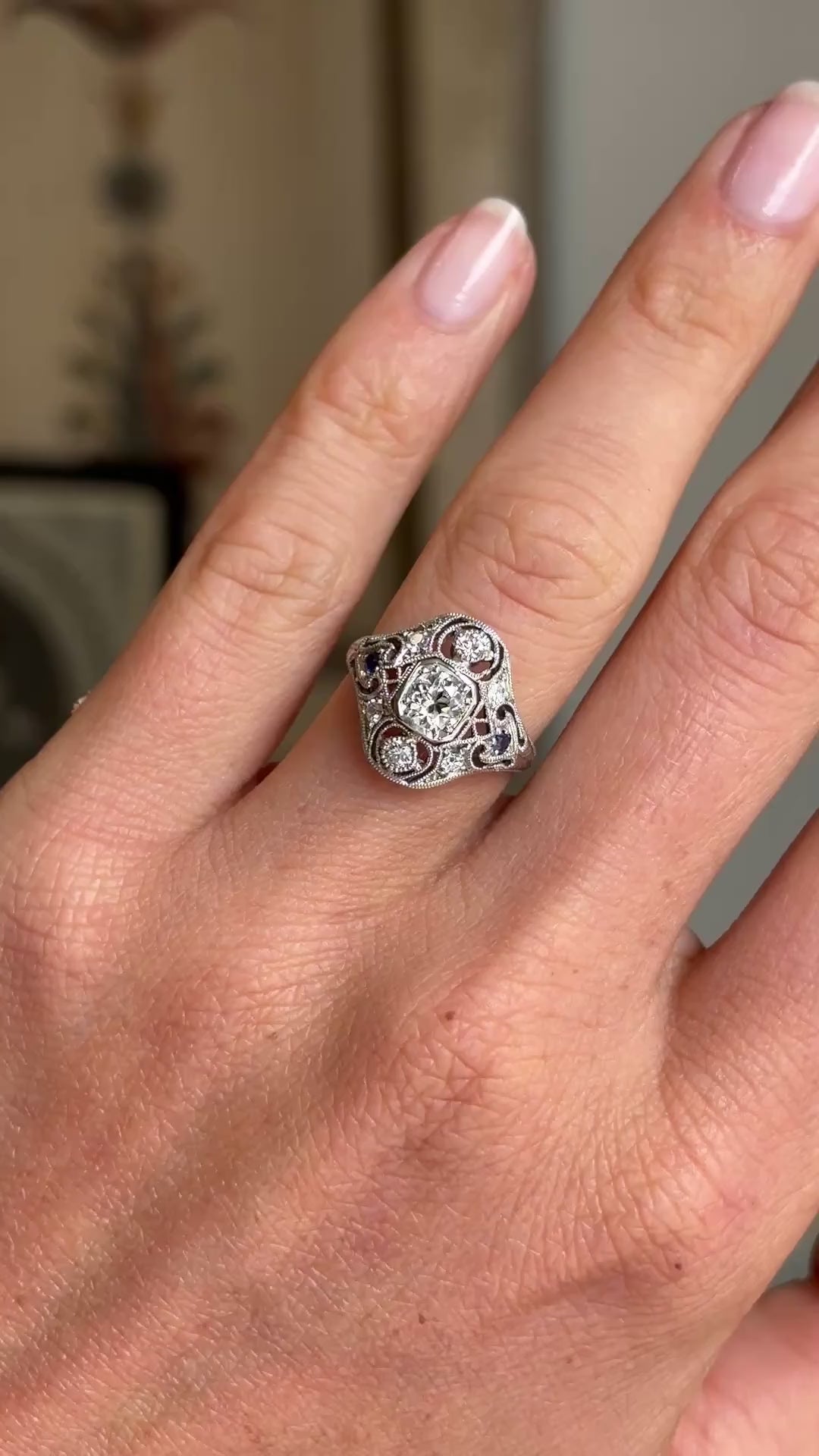 Antique Art Deco diamond and sapphire cluster ring in platinum worn on hand, showcasing a vibrant blue sapphire surrounded by sparkling old-cut diamonds, filmed against a softly lit vintage interior backdrop.