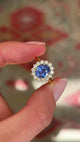 Antique-inspired intense cornflower blue sapphire and diamond cluster engagement ring featuring an oval cornflower blue sapphire encircled by brilliant-cut diamonds, held by hand for detailed viewing.