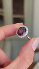 Vintage 1960s garnet and diamond cluster engagement ring featuring an oval garnet encircled by brilliant-cut diamonds, held by hand for detailed viewing.