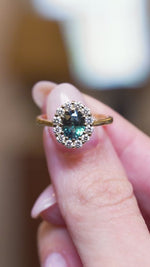 Antique inspired green sapphire and diamond cluster engagement ring held between fingers in motion.