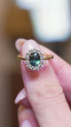 Antique inspired green sapphire and diamond cluster engagement ring held between fingers in motion.