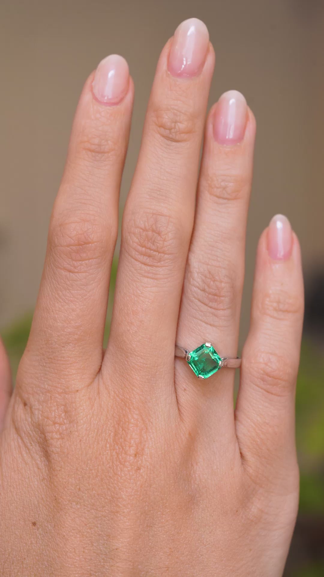 A video of a hand wearing a Vintage, 1990s emerald solitaire engagement ring on a wedding finger and rotated slowly to show the profile of the ring once worn on a finger. Against a softly blurred indoor setting. 