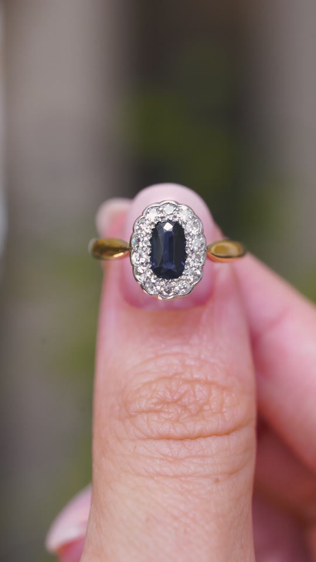 A video of an Antique, Edwardian oval-cut sapphire and diamond cluster engagement ring on a yellow gold band held between the thumb and finger and turned to showcase different angles of the ring against a blurred background.