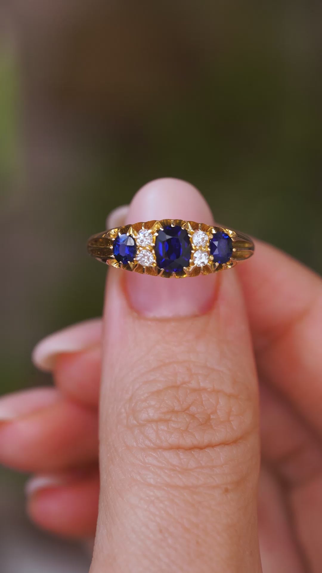A close up video of an Antique, Edwardian blue sapphire and diamond five-stone engagement ring held between two fingers and rotated slowly to show the detailed gold work of the setting and the profile of the yellow gold band. 