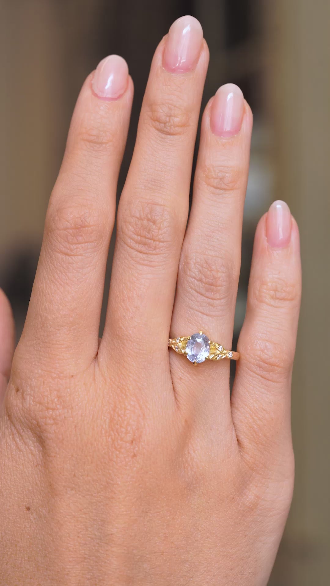 A video of a Vintage-inspired, yellow gold engagement ring set with a central pale blue oval sapphire flanked with 6 diamonds worn on a wedding finger. 