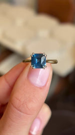 Video of an Antique-inspired, gold solitaire engagement ring held between two fingers showcasing the teal square-cut sapphire