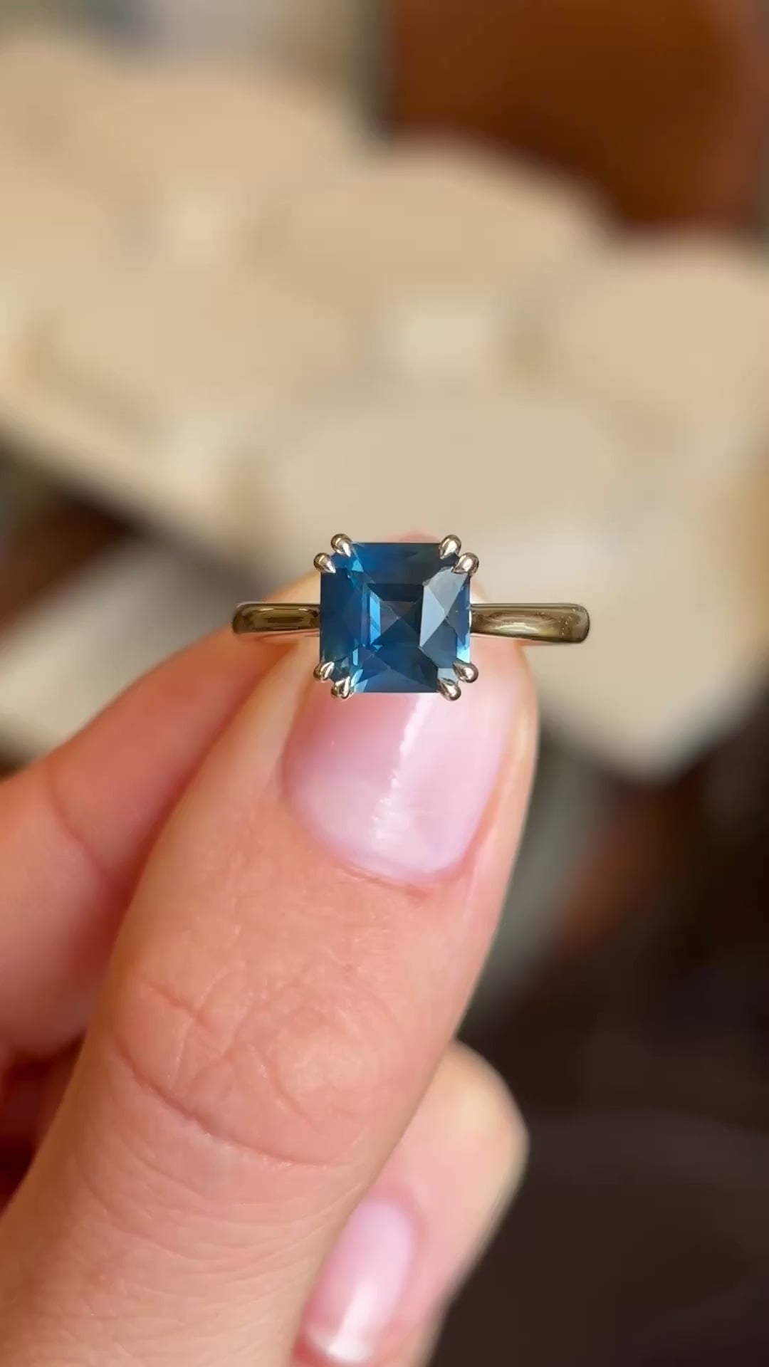 Video of an Antique-inspired, gold solitaire engagement ring held between two fingers showcasing the teal square-cut sapphire