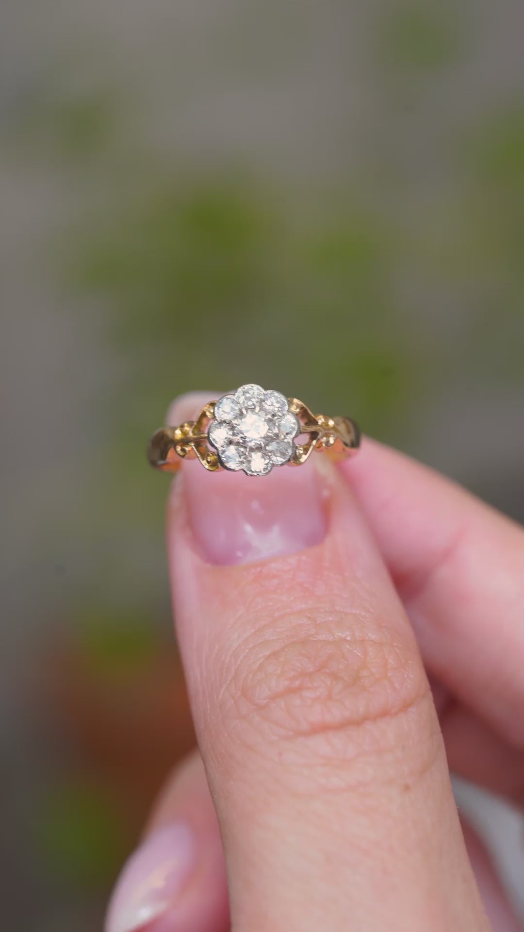 A video of an Antique, Edwardian diamond daisy cluster engagement ring from 1905 held between the finger and thumb and turned to showcase various angles and perspectives of the ring against a blurry background.