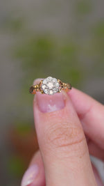 A video of an Antique, Edwardian diamond daisy cluster engagement ring from 1905 held between the finger and thumb and turned to showcase various angles and perspectives of the ring against a blurry background.